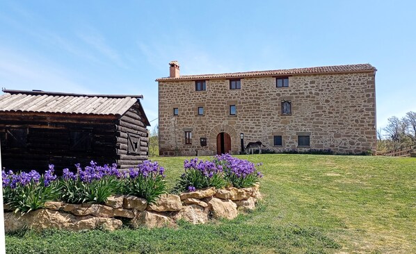A stone building with a wooden door and windows, surrounded by a grassy area with purple flowers. - Casa Rural Masia Rovira (Pinell de Solsones)
