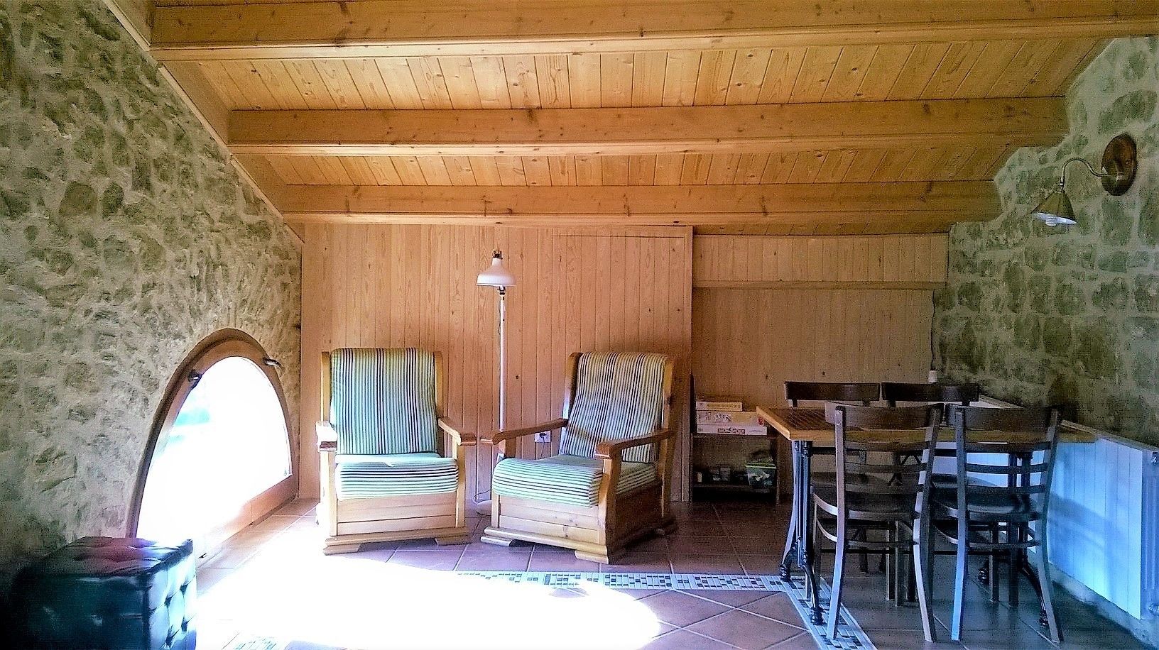 A rustic room with wooden walls and ceiling, two striped armchairs, a wooden table with chairs, and a stone wall with an arched window.