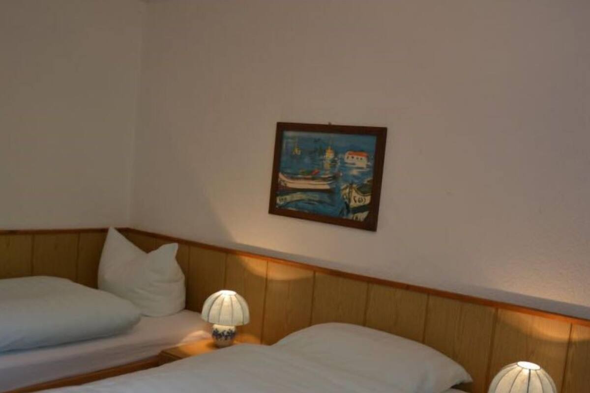 Une chambre d’hôtel avec deux lits simples, des têtes de lit en bois, du linge de lit blanc et un tableau encadré représentant un bateau, accroché au mur.