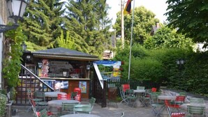 Een terras met tafels en stoelen, omringd door groen en een gebouw met een bord.