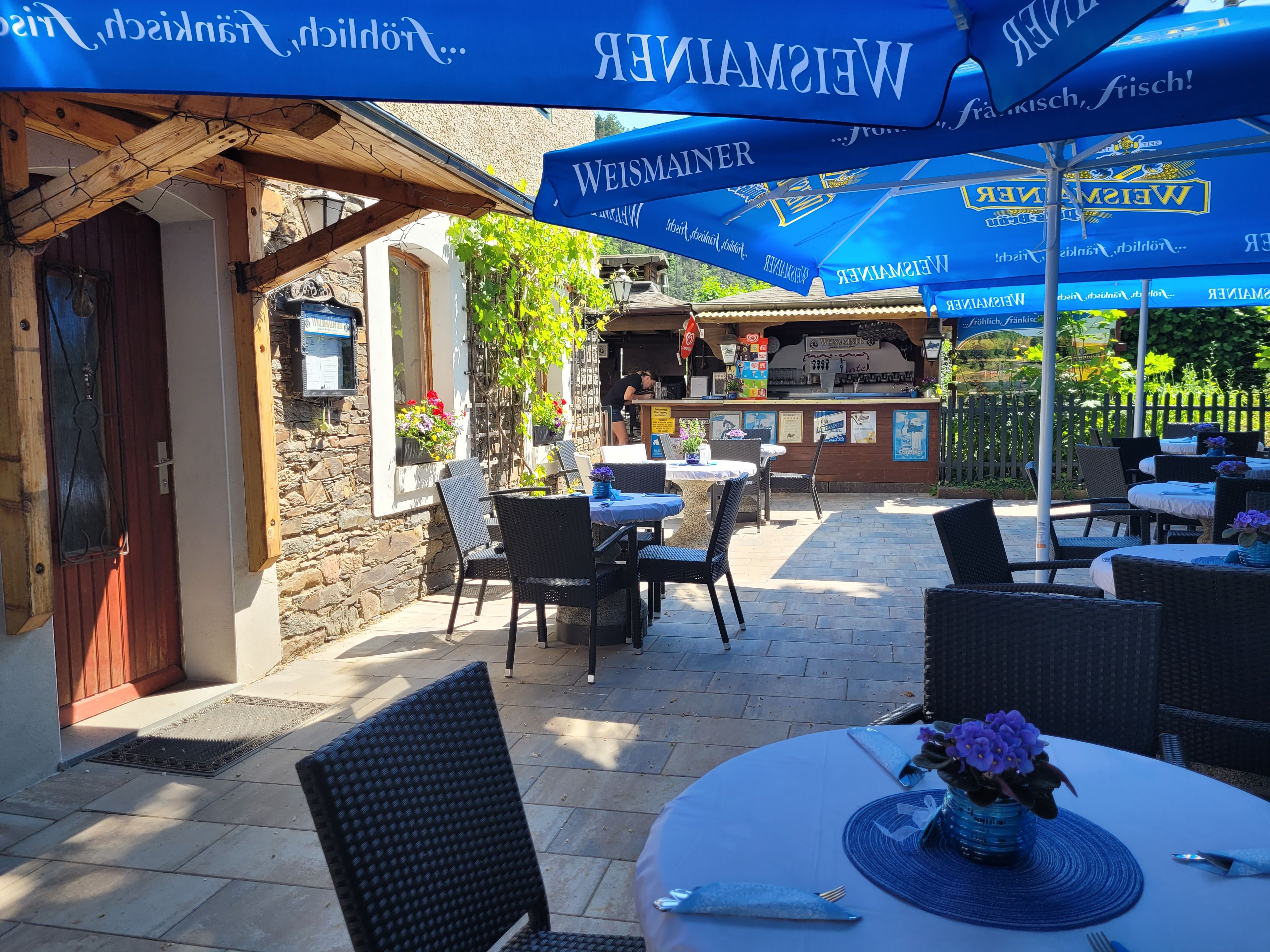Ein Außenbereich mit Sitzgelegenheiten, Tischen, Stühlen, einer Bar und Sonnenschirmen mit Branding.