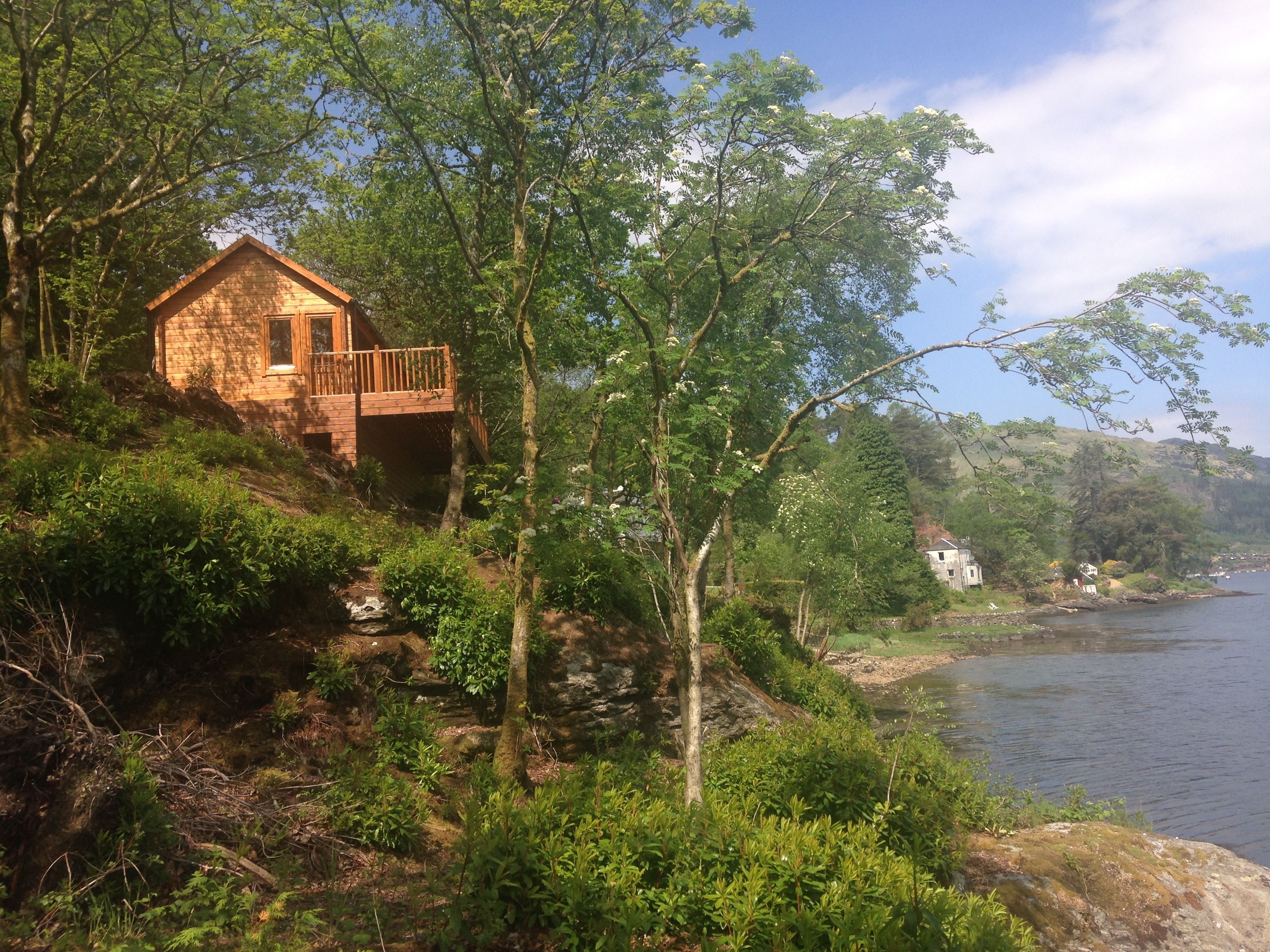 A wooden cabin with a balcony overlooking a river, surrounded by dense greenery and trees.