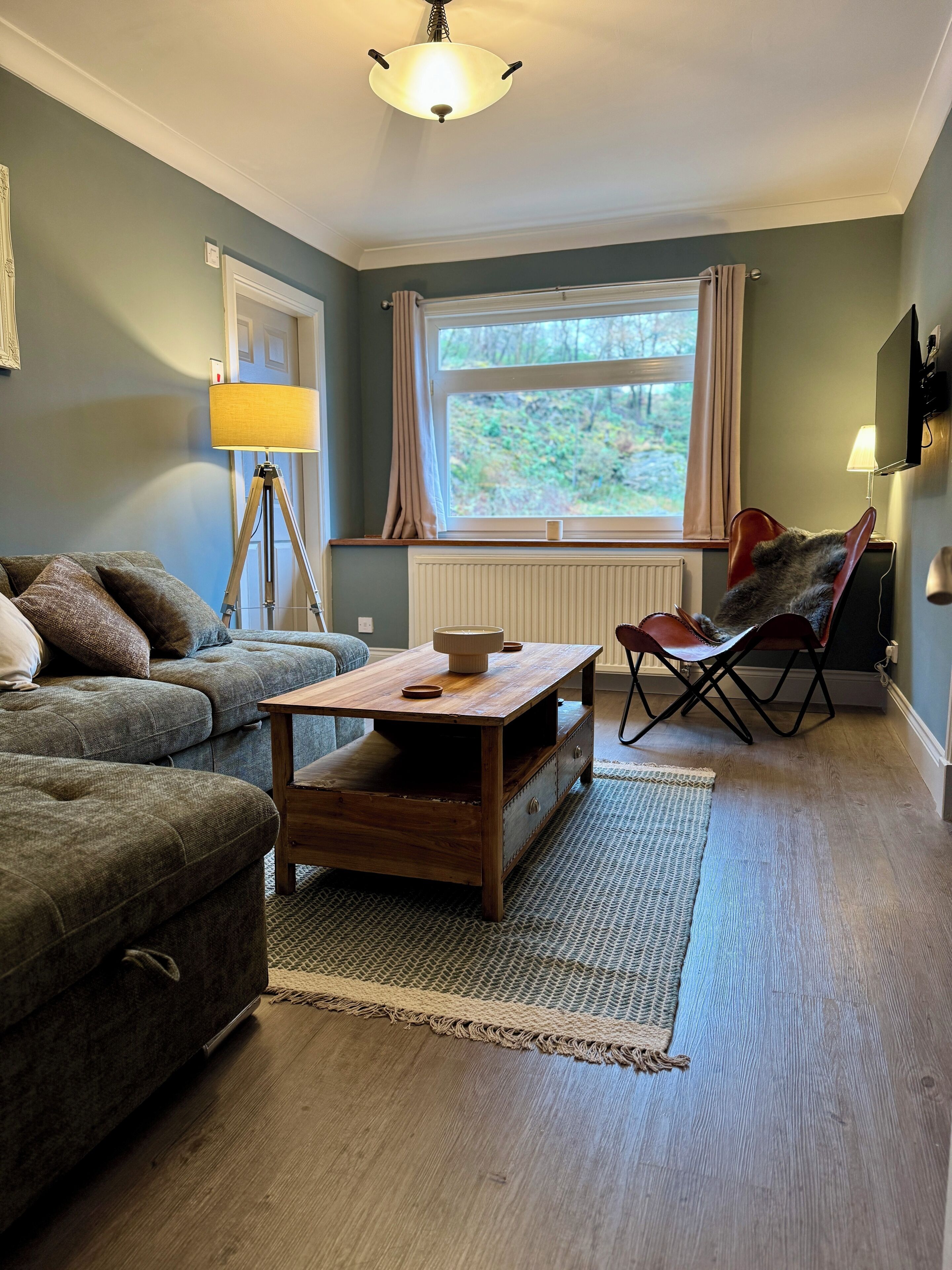 Una sala de estar acogedora con un sofá, una mesa de centro y una silla. Se ve una ventana con cortinas y vistas a zonas verdes.