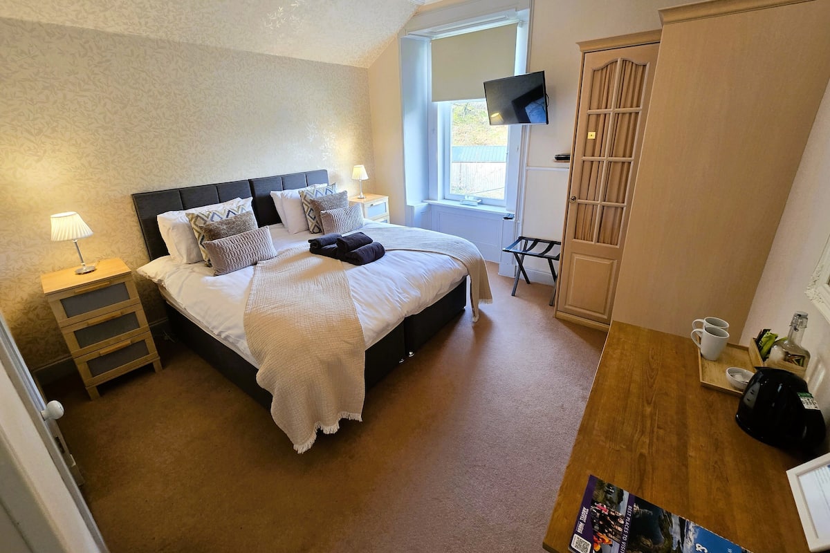 Ein ordentlich gemachtes Bett mit weißer Decke und mehreren Kissen, ein Nachttisch mit Lampe, ein kleiner Tisch mit Tassen und Wasserkocher sowie ein an der Wand befestigtes Fernsehgerät.