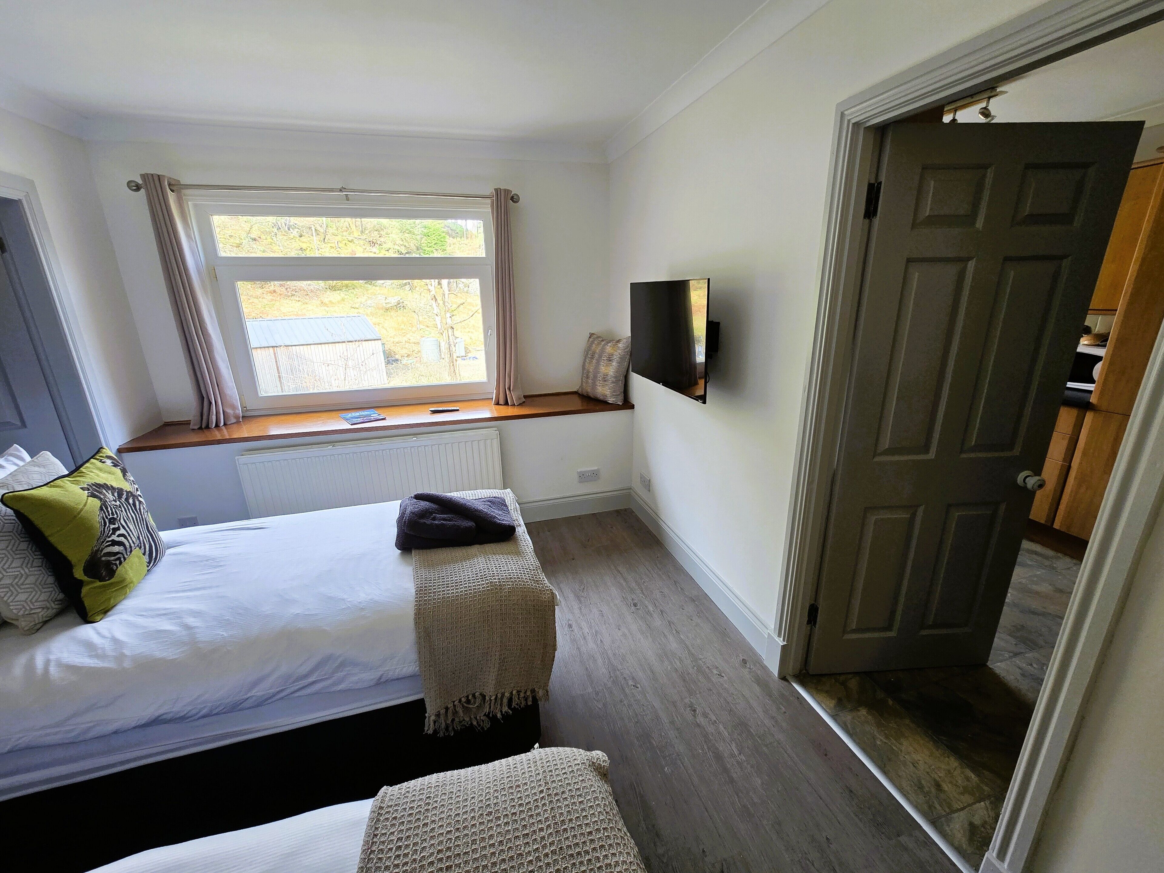 Une chambre à coucher comprenant un lit, un coin fenêtre, une télévision et une porte donnant sur une autre pièce.