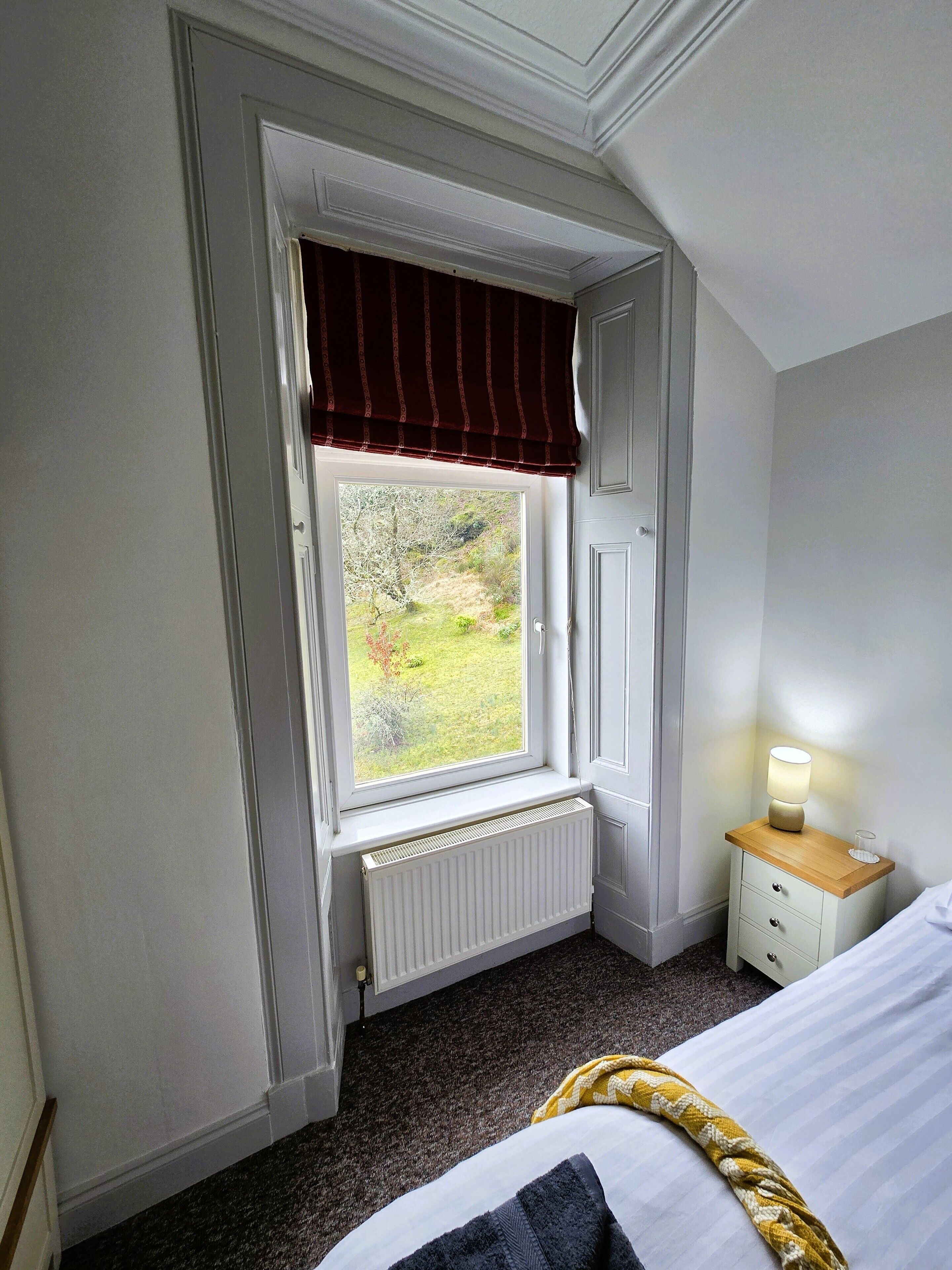 A bedroom with a window, radiator, bedside table with a lamp, and a bed.