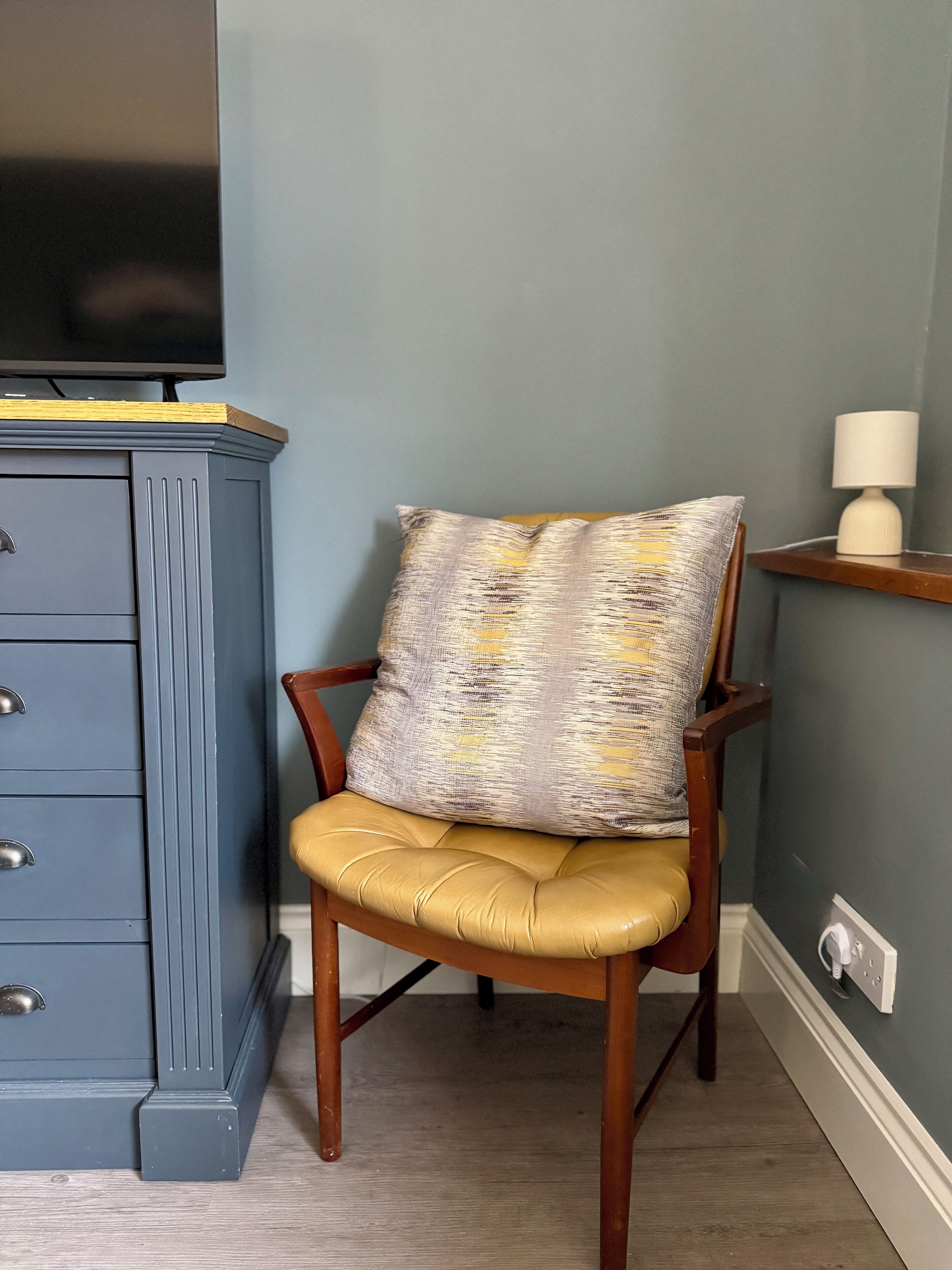 A chair with a patterned cushion and a wooden frame, positioned next to a blue cabinet with drawers and a television on top.