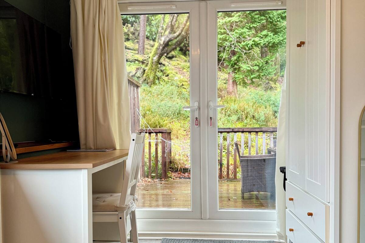 Ein Zimmer mit Schreibtisch, Stuhl, Kleiderschrank und einer Tür zu einem Außenbereich mit Holzterrasse und Grünpflanzen.