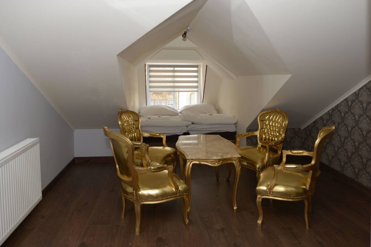 Een kamer met een schuine kap, een bed met wit beddengoed, een raam met jaloezieën en een tafel met vier stoelen.