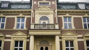 Een groot, statig gebouw met een symmetrische gevel, een centrale ingang met een decoratieve boog en meerdere ramen met luiken.
