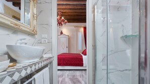 A bathroom with a large white sink, a glass shower enclosure, and a bed with red bedding. - Cà Patron (Venice)