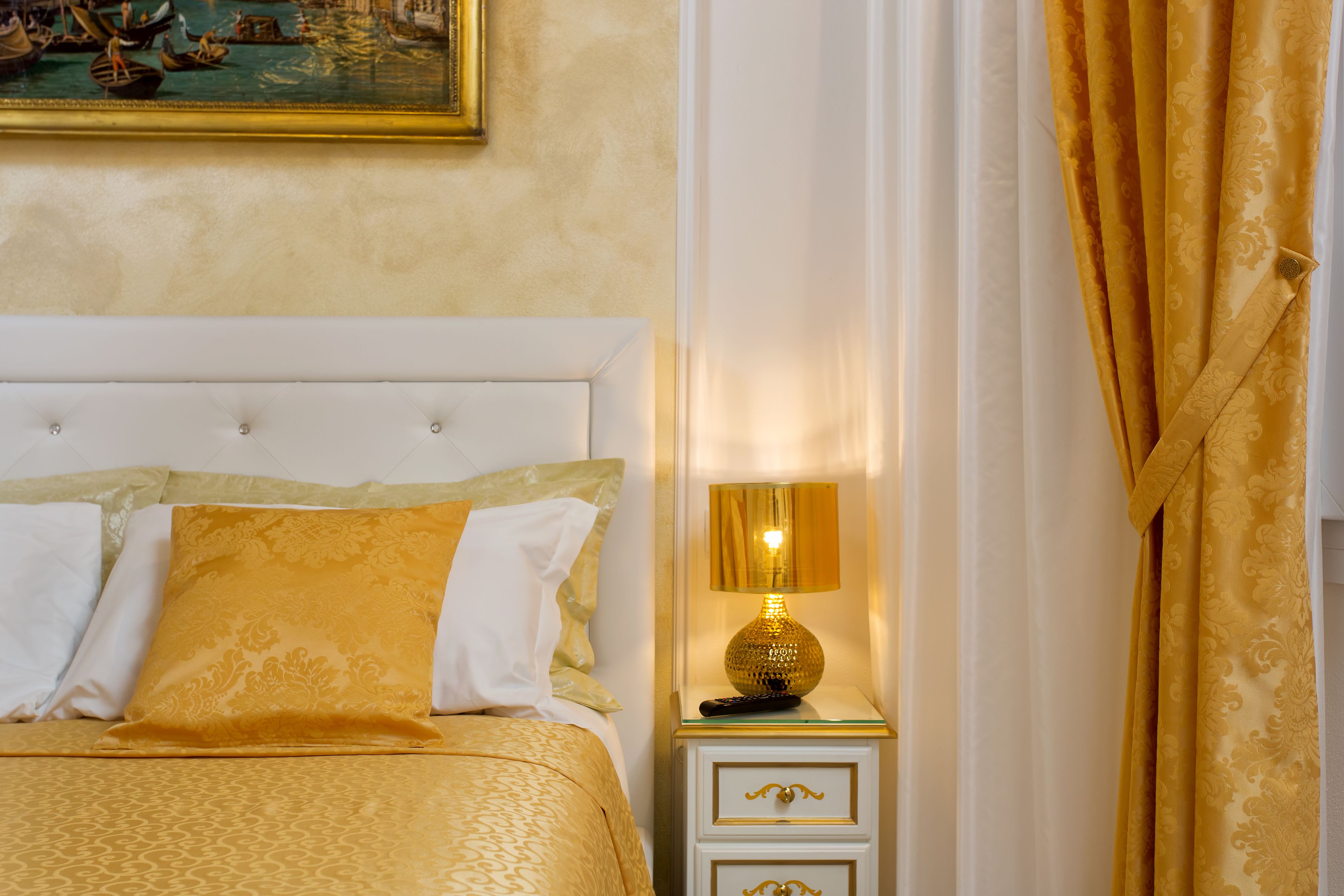 A neatly made bed with a gold and white color scheme, a bedside table with a lamp, and a painting on the wall.