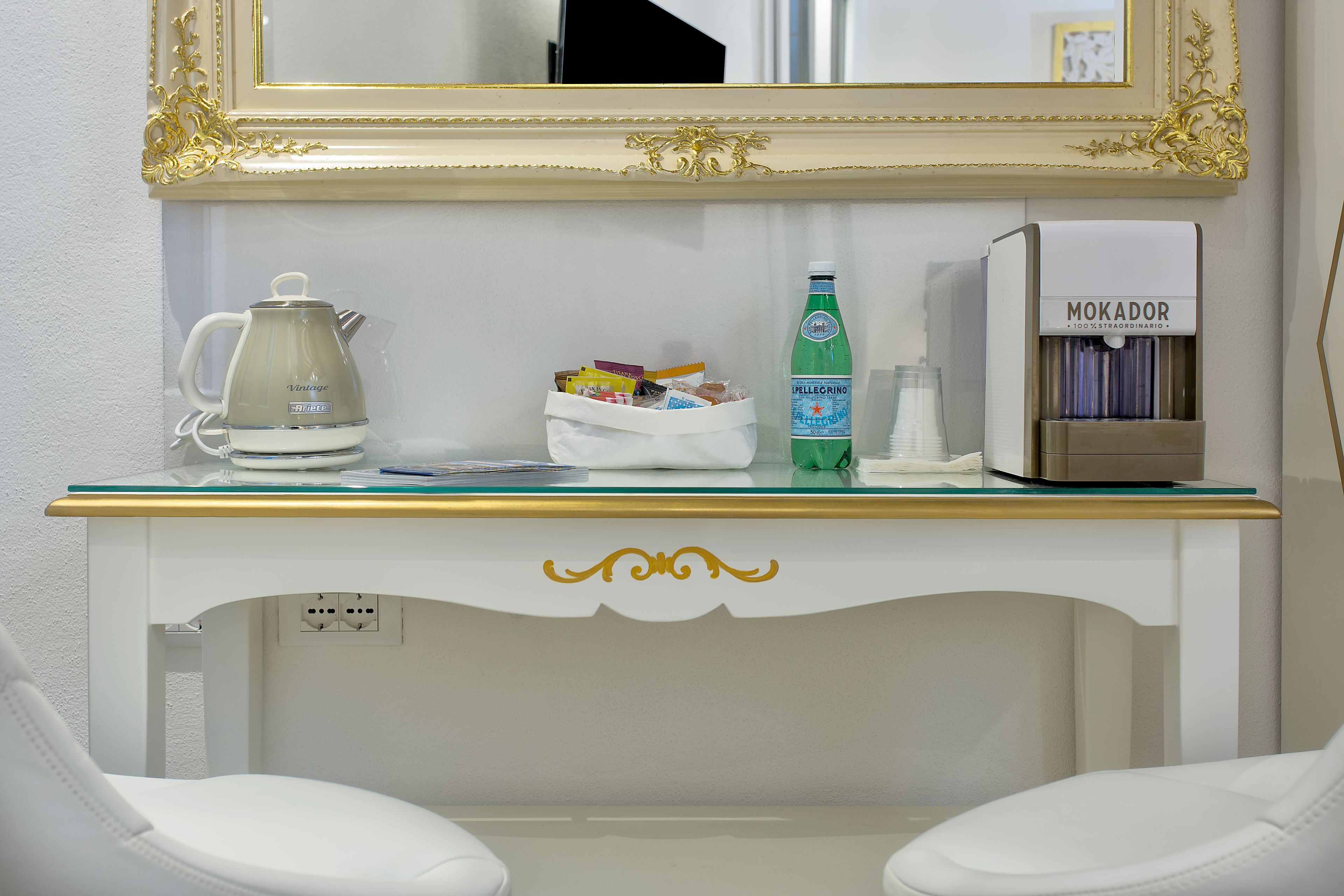A white desk with a coffee maker, a bottle of Pellegrino water, a bag of snacks, and a Mokador water dispenser.