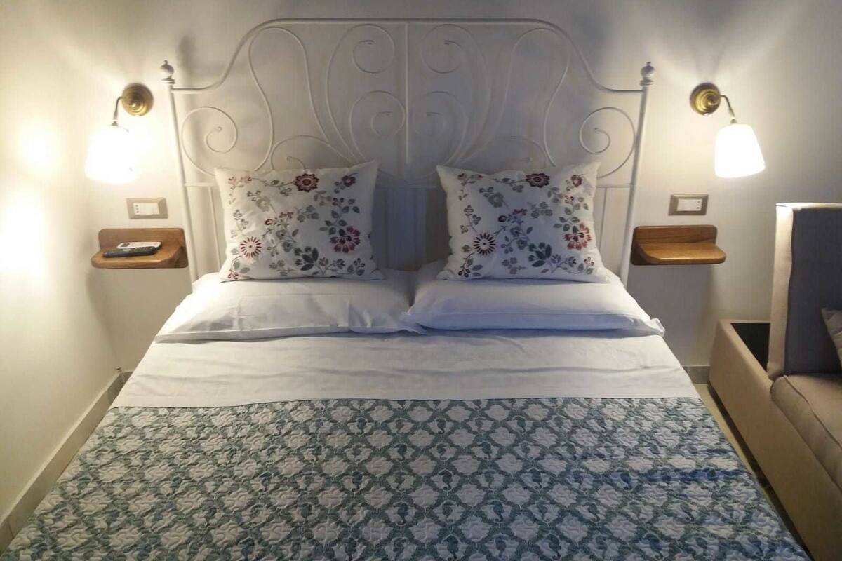 Un lit bien fait, agrémenté de coussins à motifs floraux, d’une couvre-lit à dessins et de lampes de chevet posées sur des tables de nuit en bois.