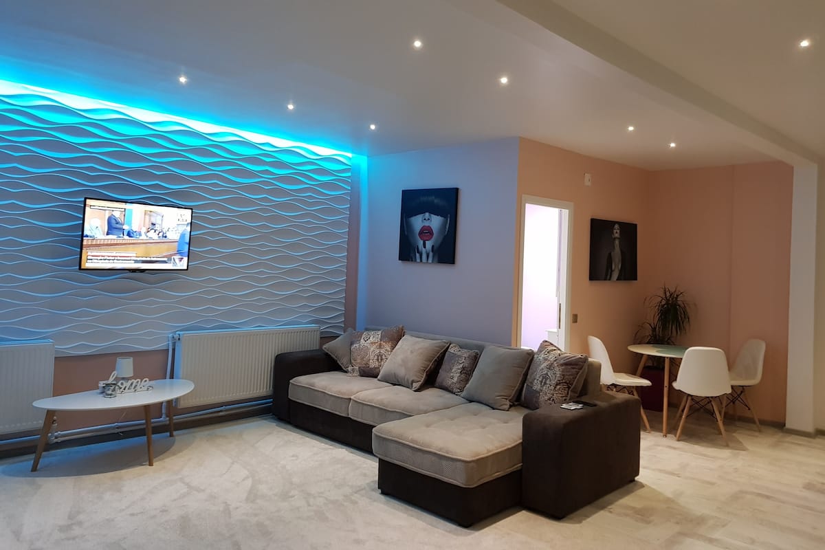 Een moderne woonkamer met een hoekbank, een kleine ronde tafel en een flatscreen-televisie aan een gestructureerde muur.