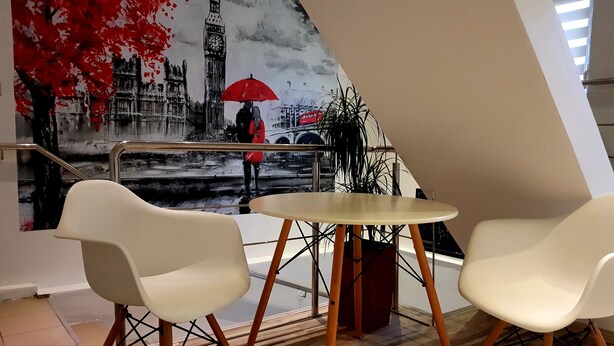 A modern interior with a wooden floor, white chairs, and a round table. A large painting of a cityscape with a red umbrella is visible in the background. - Art Boutique Central (Tirgu Jiu)