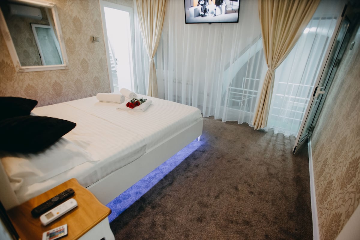 Een hotelkamer met een bed, nachtkastje met afstandsbedieningen, een aan de muur bevestigde televisie en een raam met gordijnen.