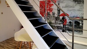 A modern staircase with a metal railing, a white chair, and a mural of a rainy street scene with a bridge and a red umbrella. - Art Boutique Central (Tirgu Jiu)