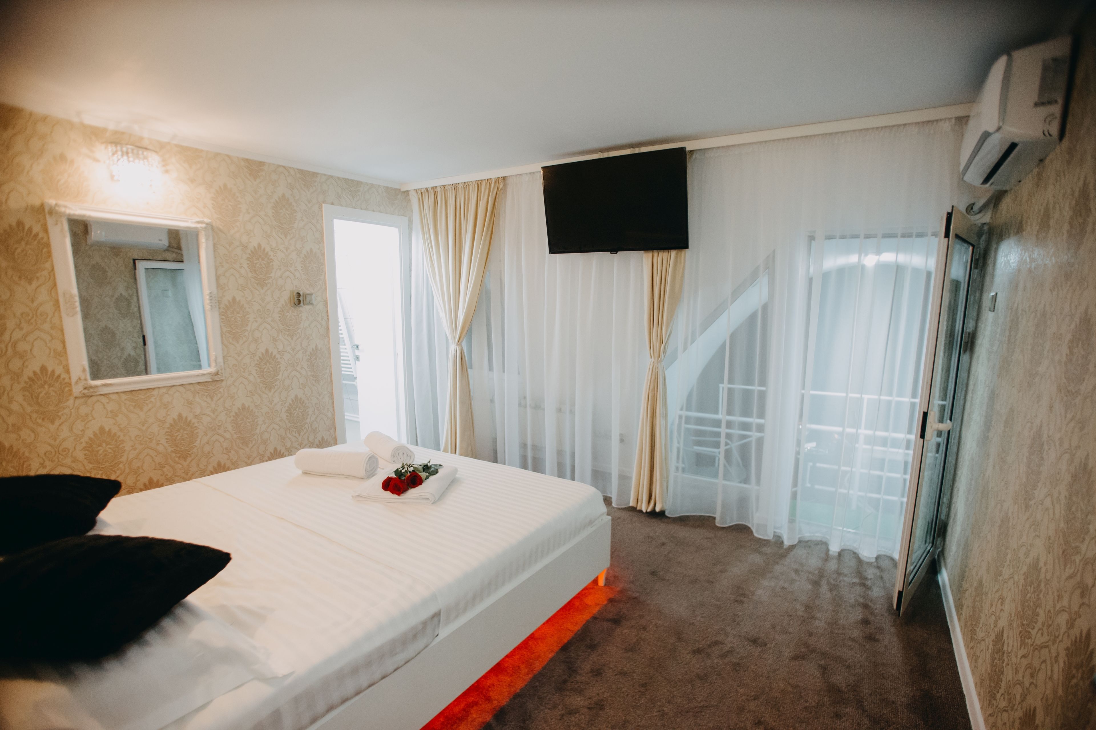 A hotel room with a bed, a flat-screen TV, a window with curtains, and a staircase visible through the window.