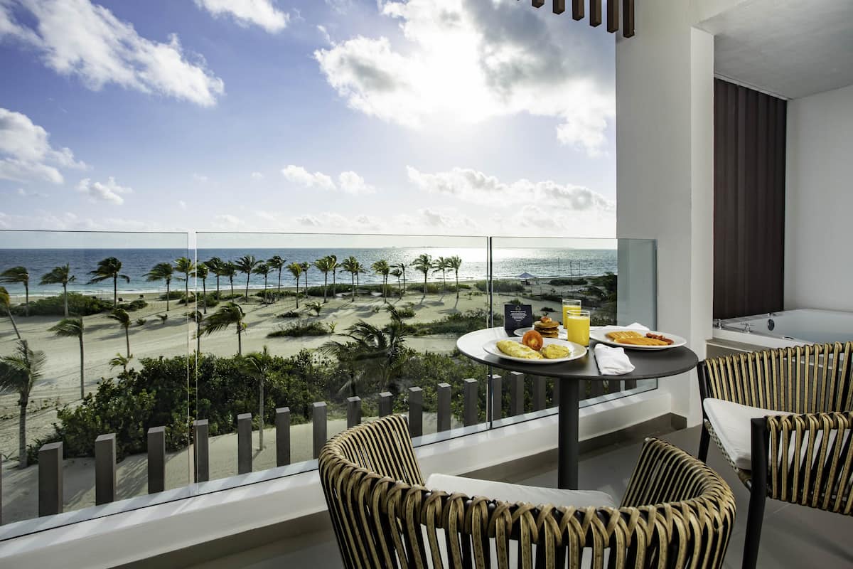 A balcony with a view of the beach, a dining table with plates and a glass of juice, and wicker chairs.