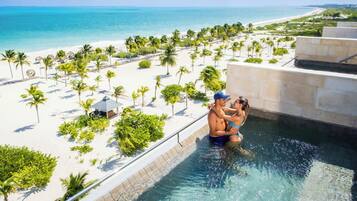 Área de piscina com vista para uma praia e oceano, cercada por palmeiras e ambiente de resort.
