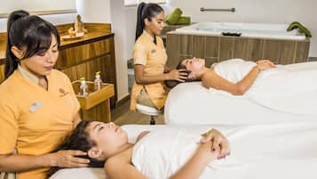 Ein Spa-Raum mit zwei Gästen, die auf Liegen liegen, einem Empfangsbereich mit Holztresen und einer großen Badewanne mit Handtuch und Zimmerpflanze.