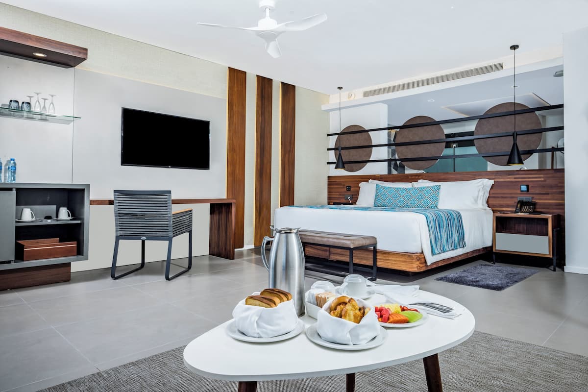 A modern hotel room with a bed, a TV, a coffee table with breakfast, and a desk with a chair.