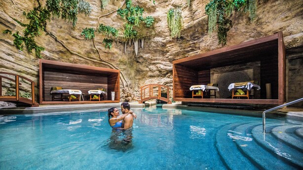 An indoor swimming pool with wooden benches and tables, surrounded by cave-like walls with greenery. - Majestic Elegance Costa Mujeres – All Inclusive (Costa Mujeres)