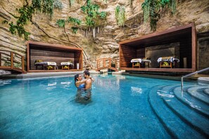 Una piscina interior con bancos y mesas de madera, rodeada de paredes similares a cuevas con vegetación.