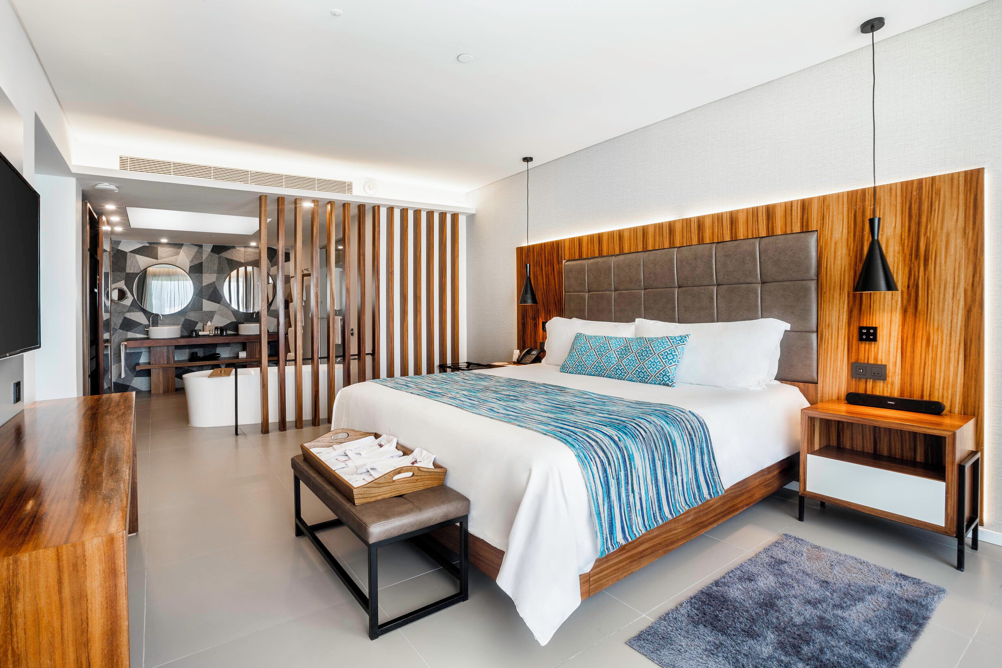 Une chambre moderne avec un grand lit, une tête de lit en bois, des tables de chevet, un banc et un téléviseur.