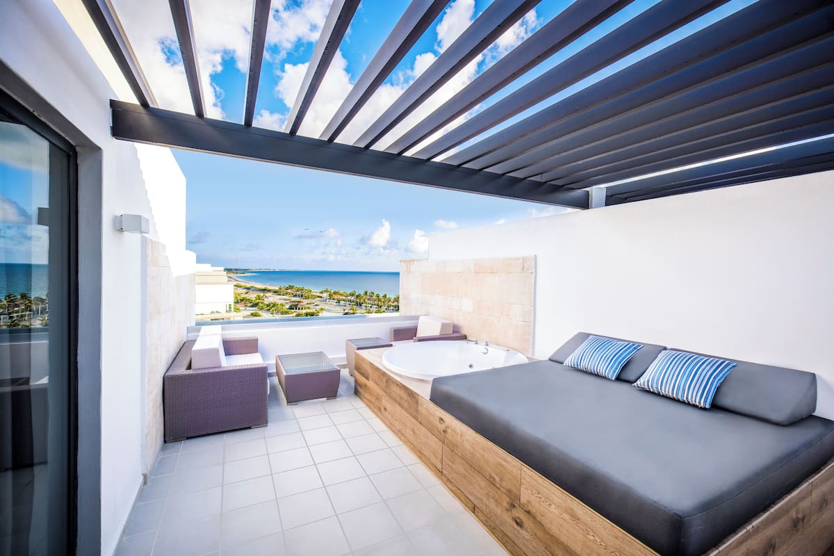 A modern outdoor balcony with a view of the ocean, a wooden bench, and a sofa with striped cushions.