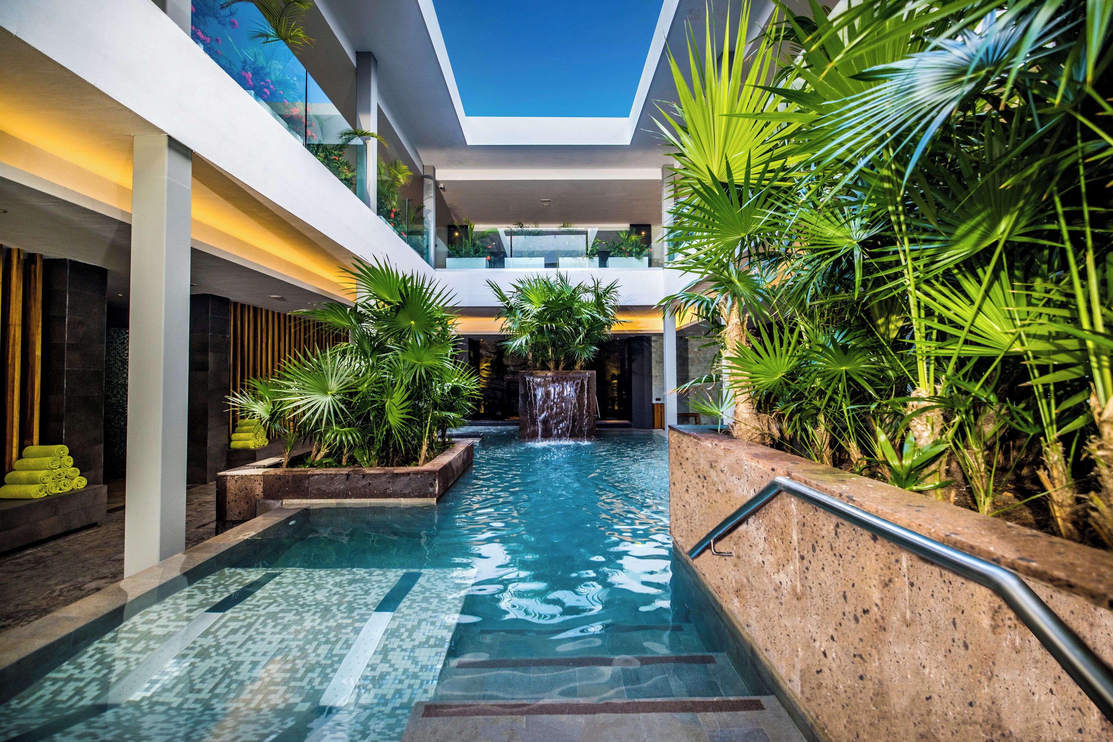 Une piscine intérieure moderne, agrémentée d’une cascade, entourée d’une végétation luxuriante et offrant une vue sur un ciel bleu dégagé grâce à une ouverture dans le plafond.