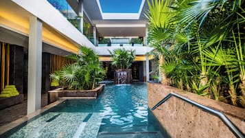 Une piscine intérieure moderne, agrémentée d’une cascade, entourée d’une végétation luxuriante et offrant une vue sur un ciel bleu dégagé grâce à une ouverture dans le plafond.