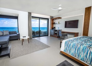 A modern hotel room with a large bed, a sofa, a small table, a desk, and a balcony with a view of the ocean.