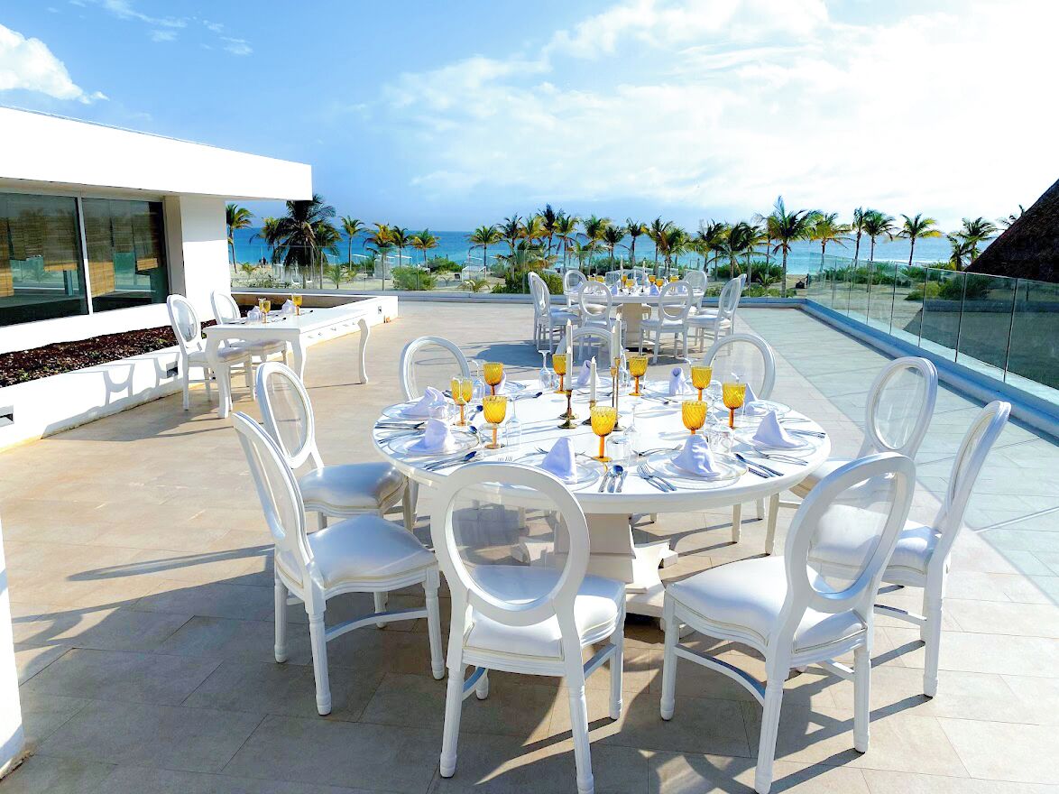Mesa redonda para exteriores con sillas blancas, un tablero de vidrio y candelabros amarillos, con vista a una playa con palmeras y cielo despejado.