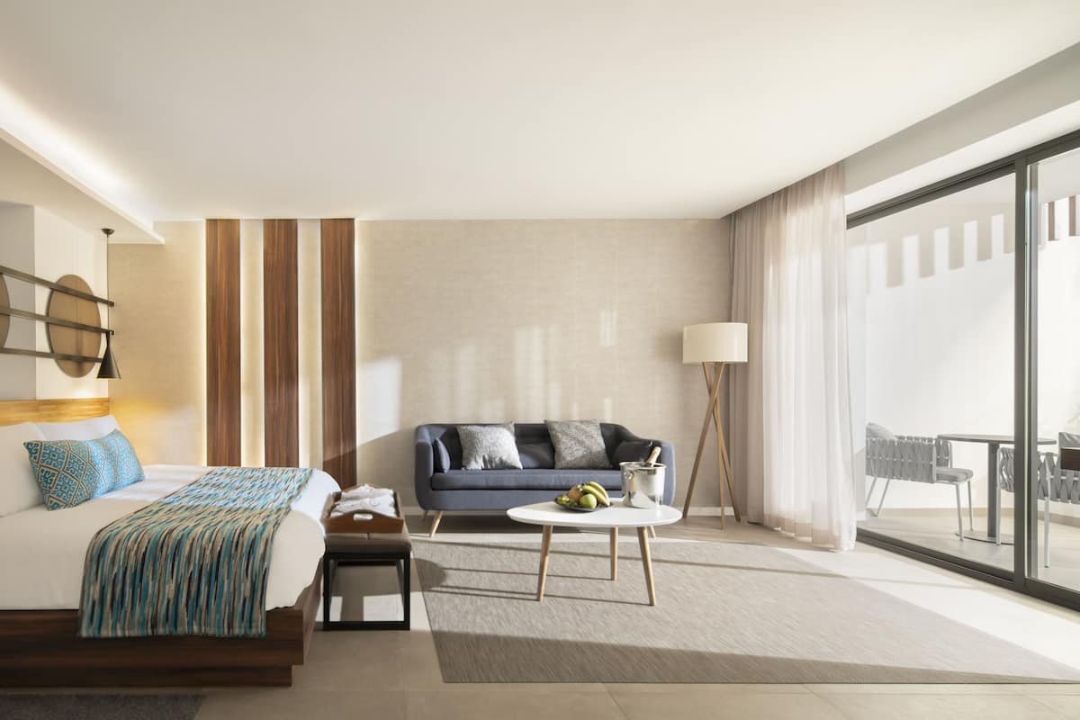 A modern hotel room with a large bed, a sofa, a small round table, and a view of the outdoors.