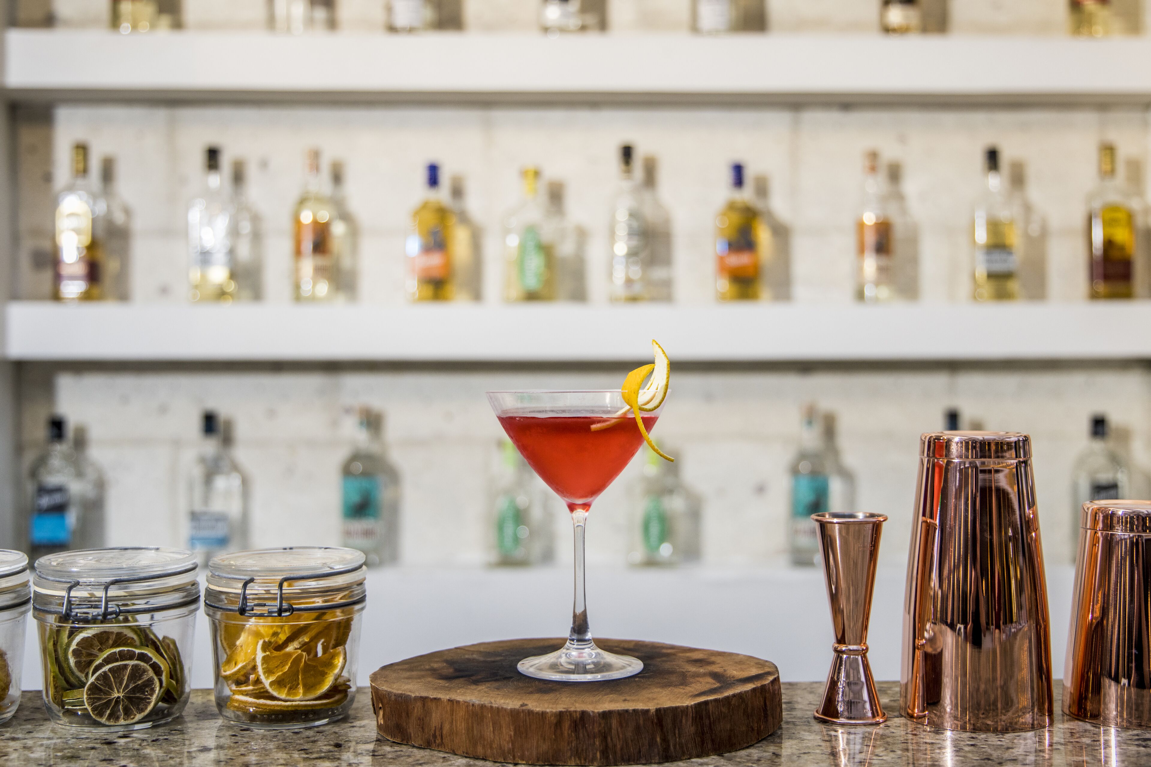 A bar area with a martini glass with a lemon twist garnish, surrounded by various bottles and jars of ingredients.