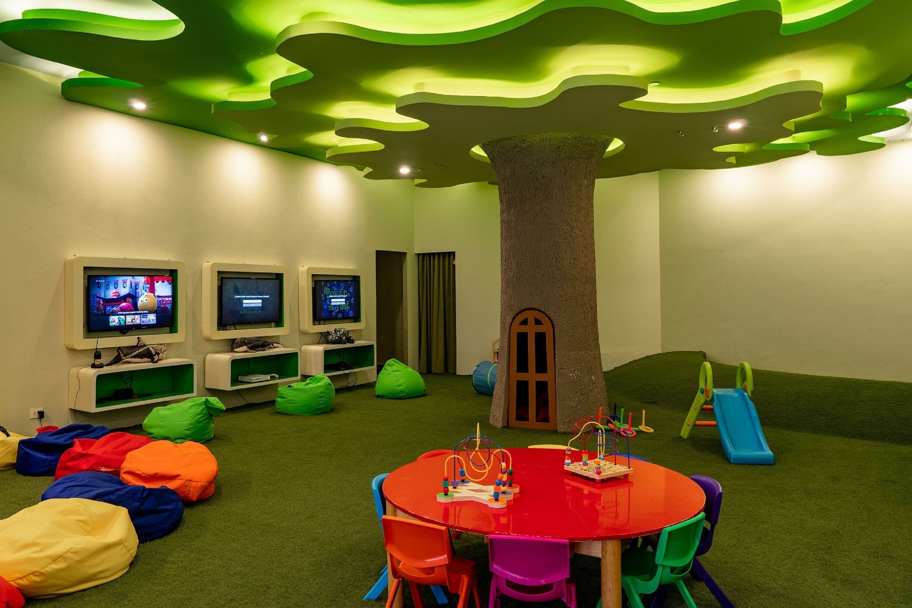 Una sala de jocs infantil amb un sostre amb forma d'arbre, una taula vermella, butaques de gresol de colors i una tobogan.