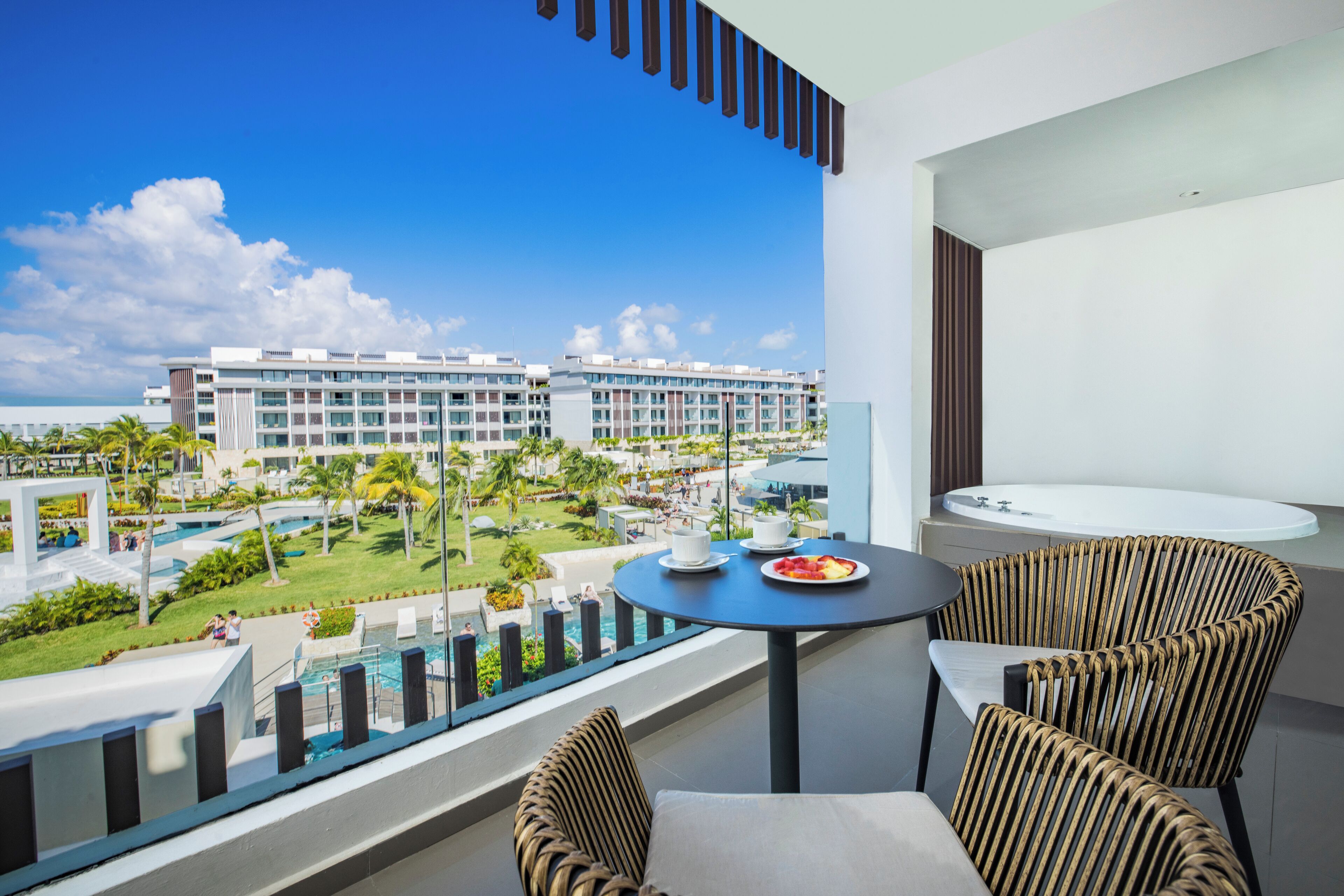 Un balcón con vista a una piscina y edificios de un resort. Hay sillas de mimbre y una mesa con un plato de comida.