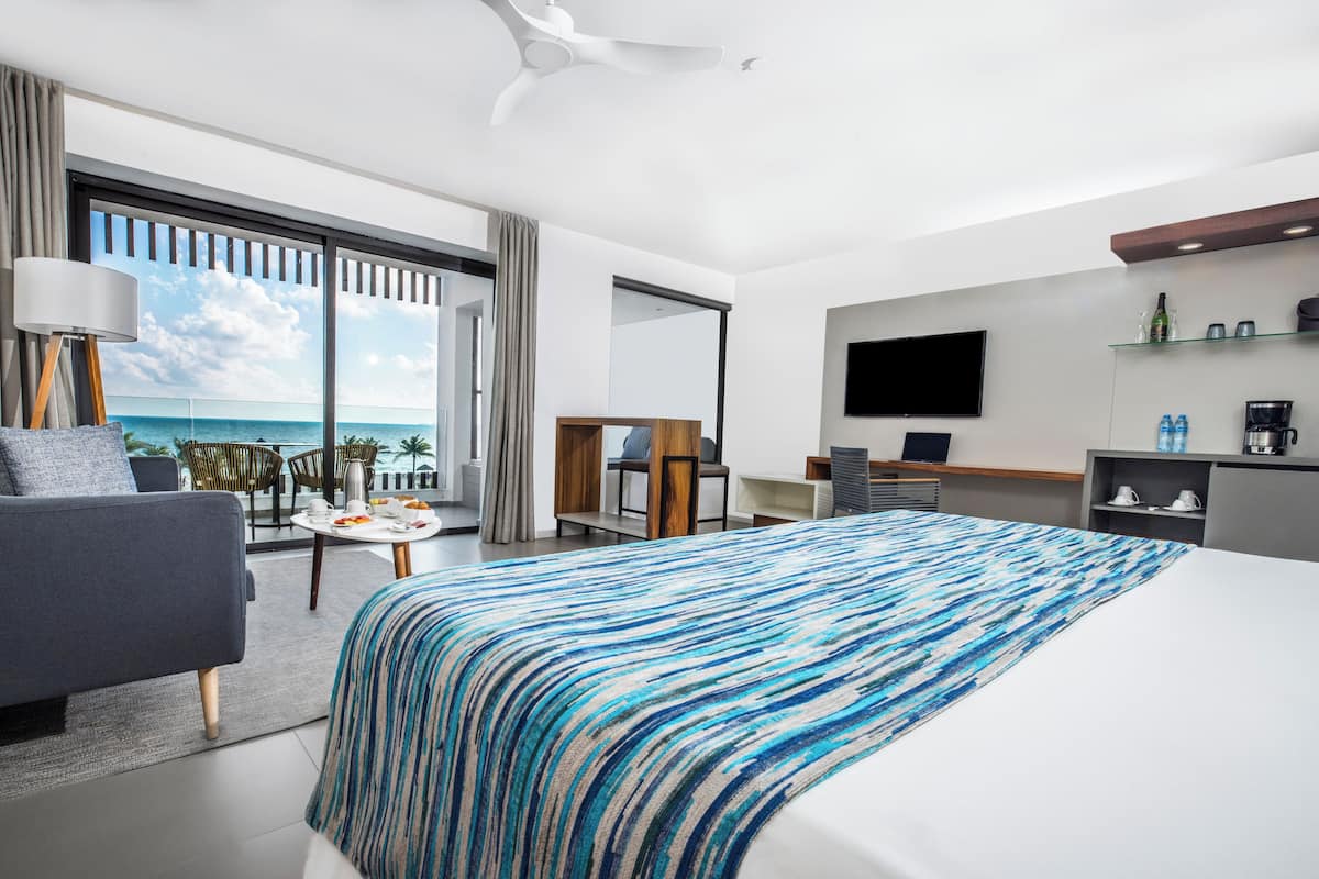A modern hotel room with a large bed, a desk, a TV, and a balcony with a view of the ocean.