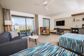 A modern hotel room with a sofa, two beds with striped bedding, a dining table, a TV, and a balcony with a city view.
