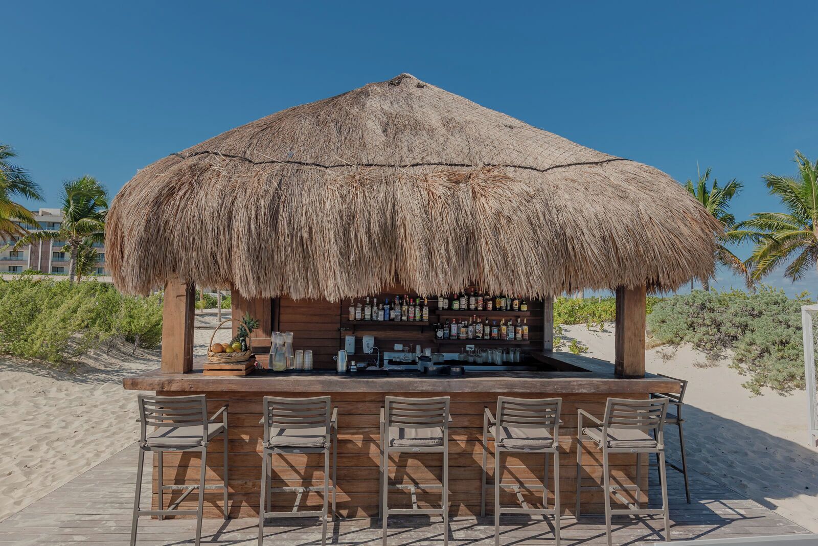 Un bar de playa con techo de paja, mostrador de bar de madera y cuatro sillas de metal.