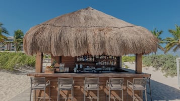 Un bar de playa con techo de paja, mostrador de bar de madera y cuatro sillas de metal.