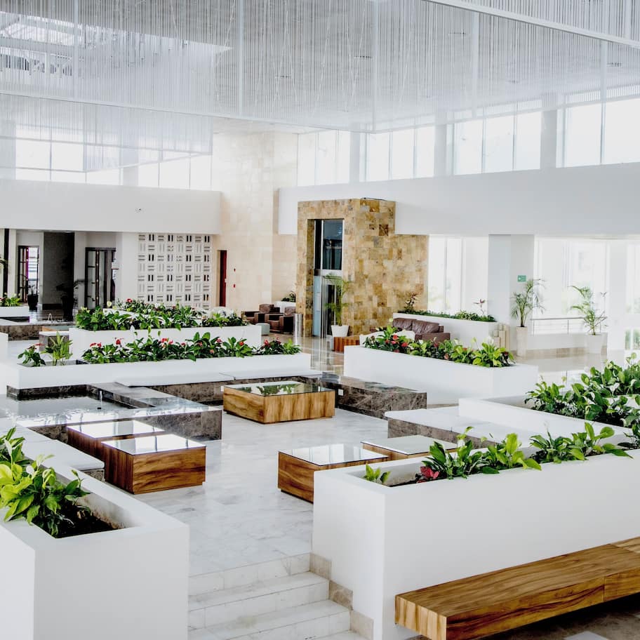 A spacious lobby with white seating areas, planters with greenery, and a modern architectural design.