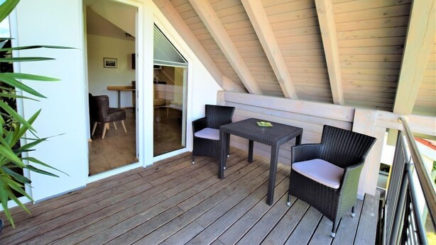 A wooden deck with two wicker chairs and a small table, a sliding glass door leading to an interior room, and a sloped ceiling. - Gästehaus Jäger (Gersfeld)