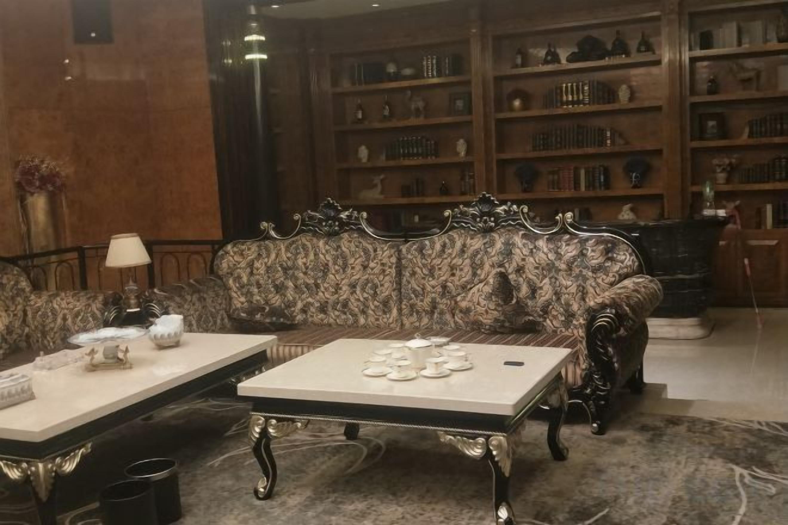 A room with a patterned sofa, two coffee tables, and a bookshelf filled with books and decorative items.