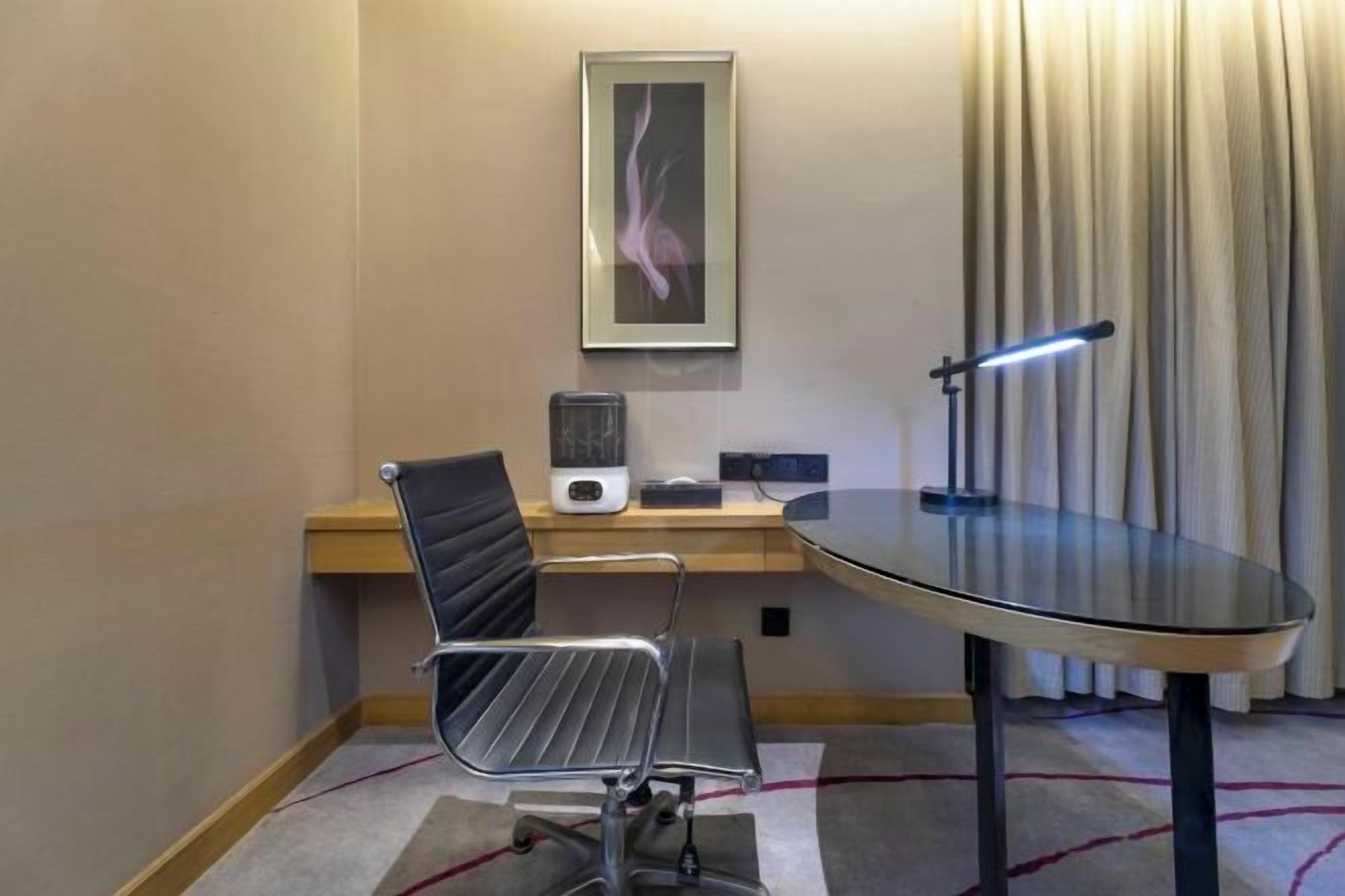 Hotellihuone, jossa on lasipöytä, musta toimistotuoli, tietokoneella varustettu työpöytä, seinällä oleva kehystetty kuva ja ikkuna, jossa on verhot.