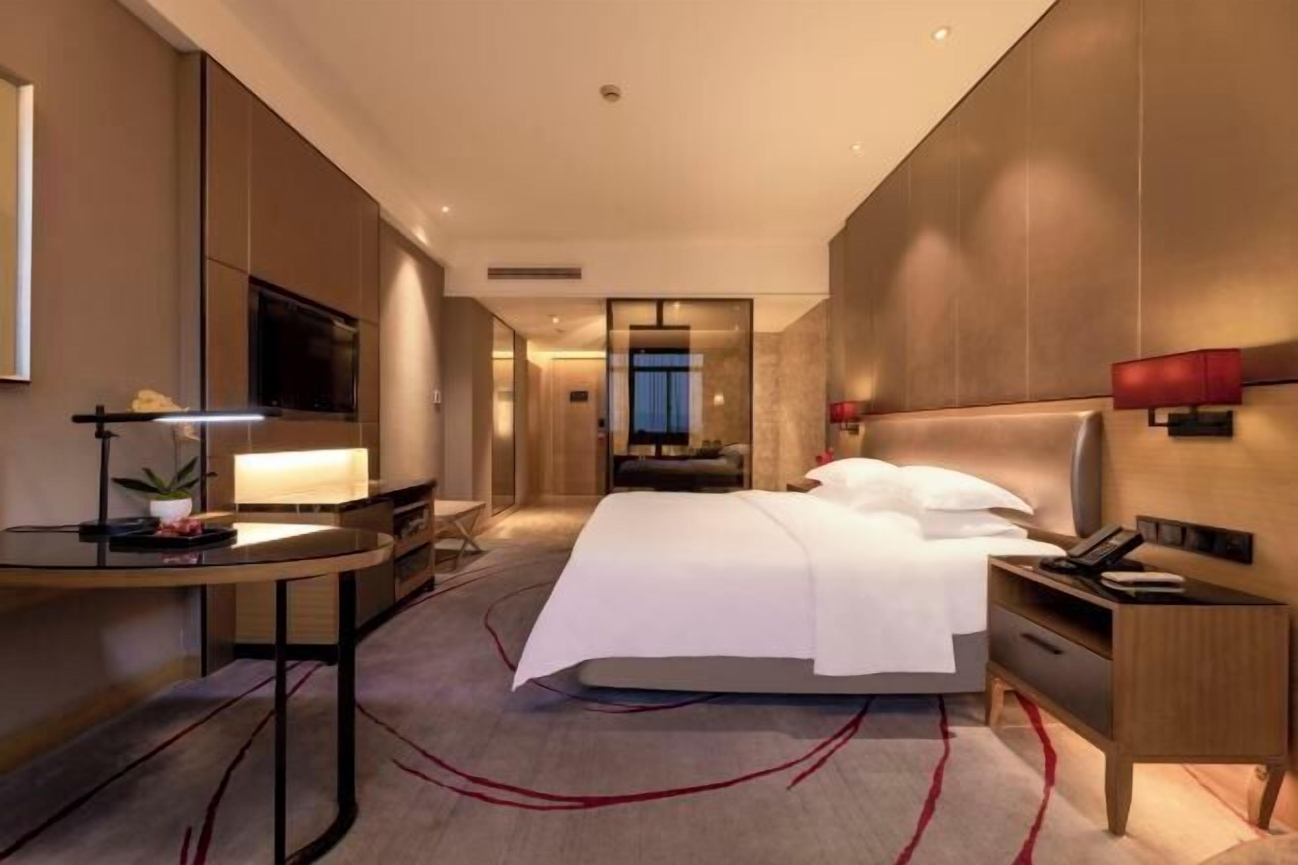 Ein modernes Hotelzimmer mit einem großen Bett, einem Schreibtisch mit Lampe, einem Fernseher und Blick auf die Stadt.