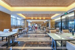 A spacious hotel lobby with long tables, comfortable seating, and a view through the glass wall.