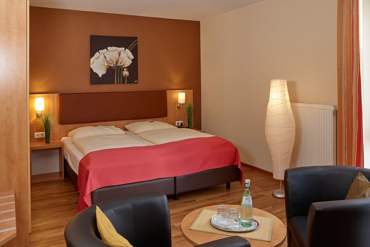A hotel room with a double bed, two bedside tables with lamps, a large vase, a sofa, and a small table with a bottle and glasses.